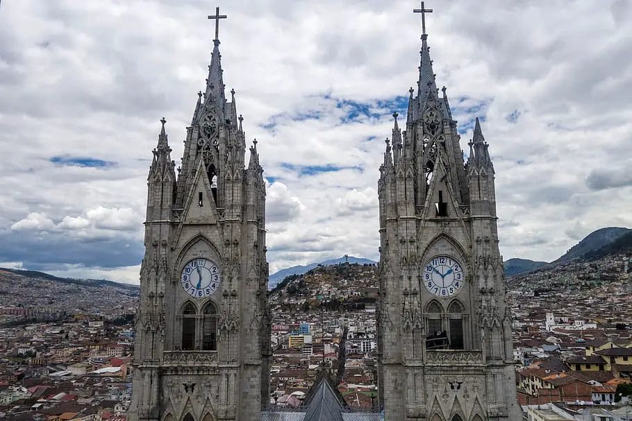 a two towers with clocks on each side with Quito in the background