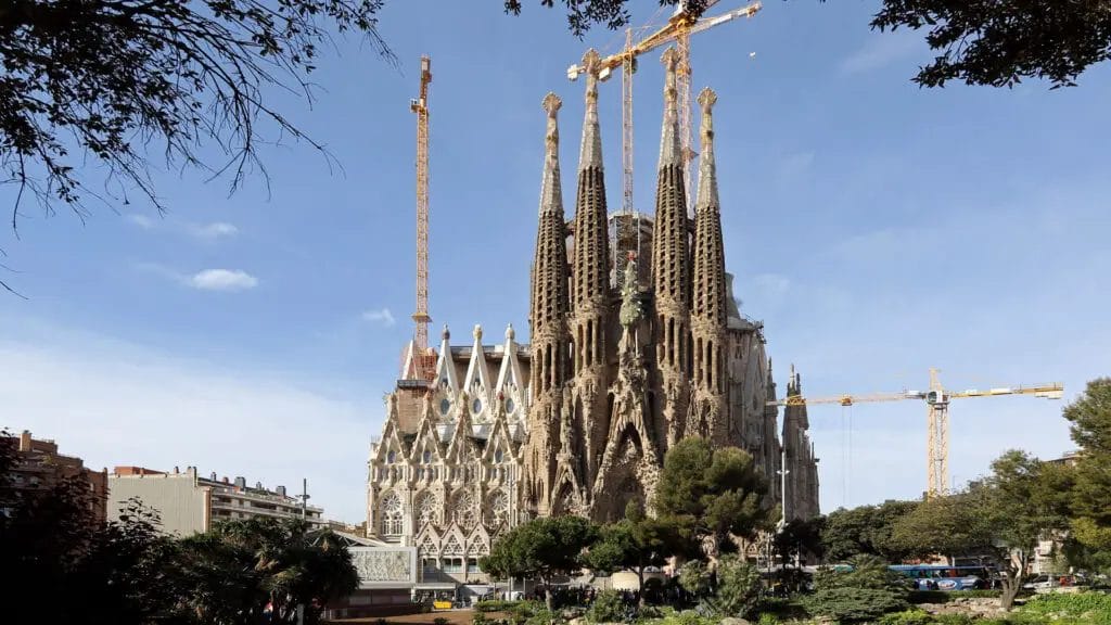A Large Building With Towers And Cranes With Sagrada Família In The Background
