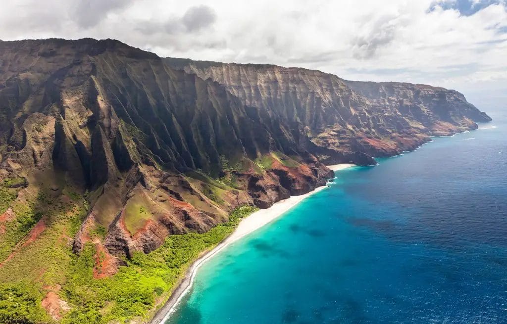 A Large Rocky Cliff With A Beach And Blue Water With Nā Pali Coast State Park In The Background
