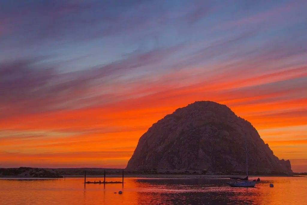a boat in the water with a mountain in the background in Morro Rock Sunset.